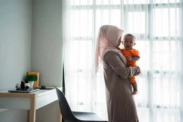 Мать азиатского мусульманина носит своего маленького мальчика, когда играет у окна — стоковое фото