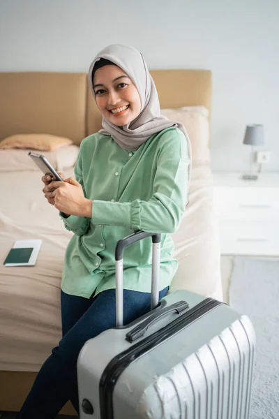 Junge asiatische Frau sitzt auf dem Bett und hält ihr Handy und ihren Koffer — Stockfoto