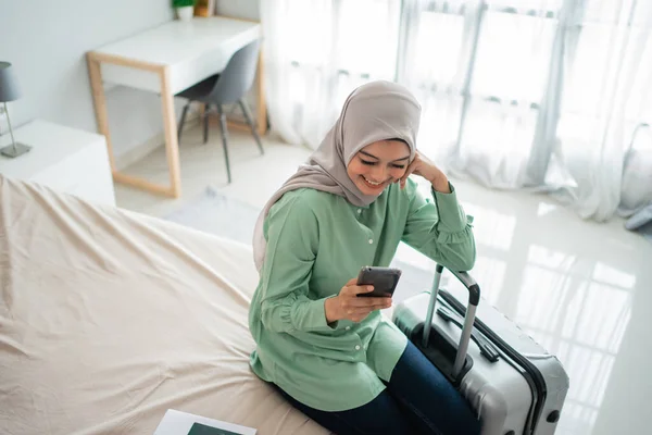Junge asiatische Frau sitzt auf dem Bett und hält ihr Handy und ihren Koffer — Stockfoto