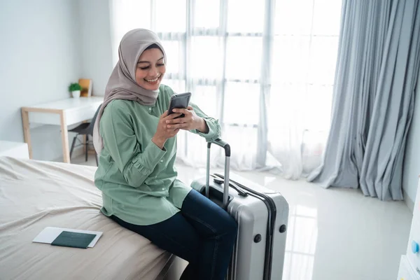 Fiatal ázsiai nő ül az ágyon kezében a mobiltelefon és bőrönd — Stock Fotó