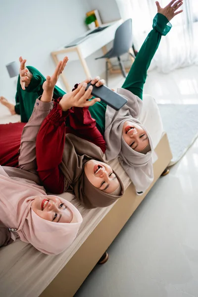 Wanita dan teman hijab asia berbaring dan mengangkat tangan di tempat tidur sambil berfoto selfie bersama — Stok Foto