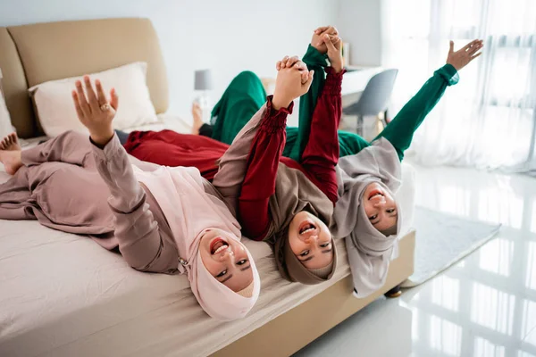 Perempuan dan teman-teman hijab asia berbaring dan mengangkat tangan di tempat tidur sambil bersenang-senang bersama — Stok Foto