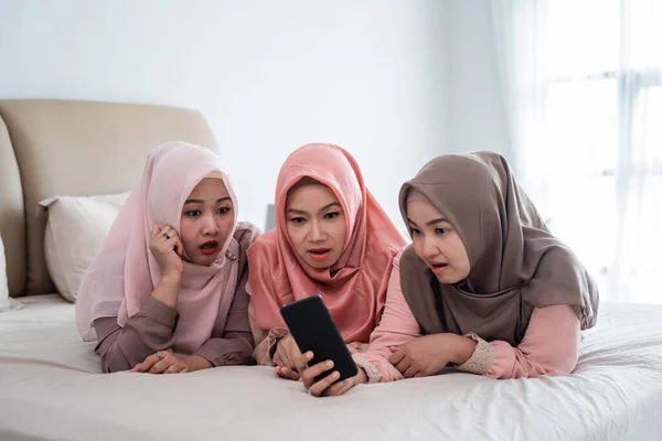 Tres hijab mujer acostada en la cama estupefacta mirando teléfono inteligente — Foto de Stock