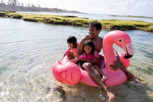 Vater und zwei Töchter reiten auf Flamingo-Boje am Strand — Stockfoto