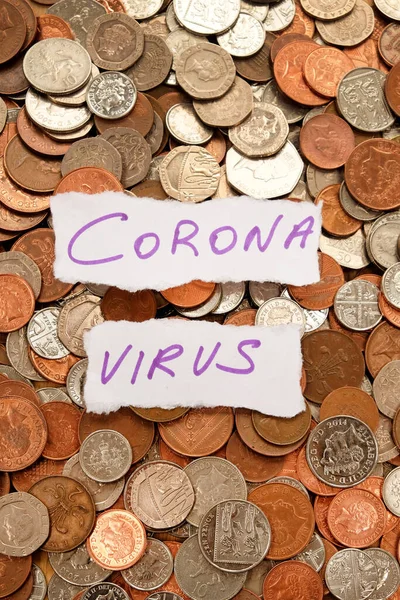 Yüzlerce gümüş ve bakır madeni paranın üzerinde duran iki parça yırtık beyaz kağıda mor mürekkeple yazılmış Corona virüsü, İngiliz parası, korona virüsü krizi ve parayı sembolize ediyor.