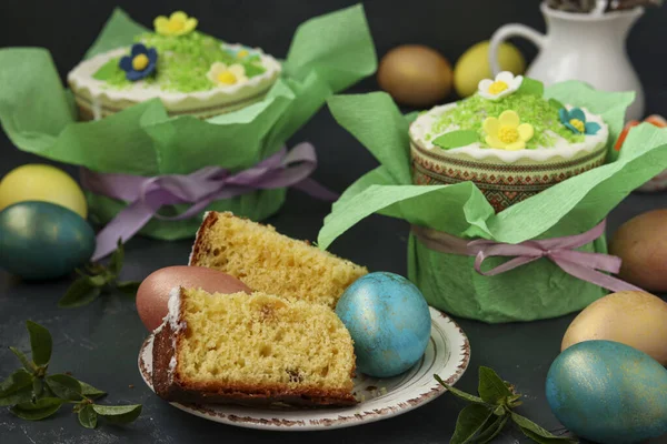 Still life with Easter cakes in gift paper and multi-colored Easter eggs on a dark background, in the foreground cut pieces of Easter cake — Stock Photo, Image