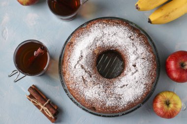 Ortasında elmalar, muzlar ve tarçınlı kek, açık mavi arkaplanda krema şekeri serpiştirilmiş, iki fincan çay, üst manzara.