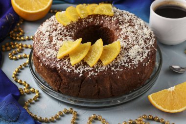 Ortasında delik olan portakallı ev yapımı kek, açık mavi arka planda hindistan cevizi taneleri ve bir fincan kahve serpiştirilmiş.