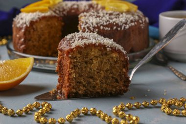 Açık mavi arka planda hindistan cevizi taneleri serpiştirilmiş portakallı ev yapımı kek ve bir fincan kahve, ön planda bir parça kek.