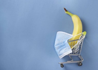 Mavi arka planda muz maskeli market arabası Coronavirus salgını sırasında alışveriş yapma tehlikesi kavramı COVID-19, kopyalama alanı.