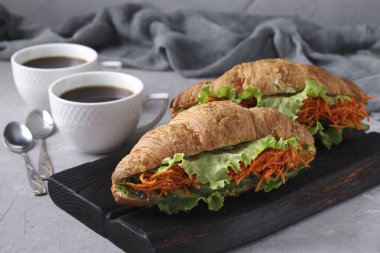 İki vejetaryen kruvasan sandviçi, marul, havuç, salatalık ve açık gri beton arka planda iki fincan kahve. Kahvaltı veya öğle yemeği konsepti. Yakın çekim.