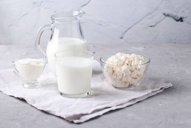 Süt ürünleri: süt, kefir veya ayran, süzme peynir ve ekşi krema şeffaf bir kase içinde, sürahi ve cam gri arka planda, yatay format