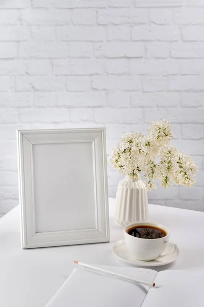 Still life with white objects on a white background. Blank photo frame, diary, notebook, pencil, vase with flowers and a cup of coffee. Layout for the morning concept.