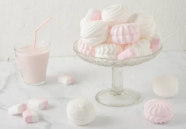 White and pink marshmallows in a glass vase on a white table. There\'s a glass mug of pink yogurt next to it. Soft light in high key.