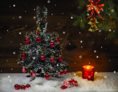 Üzerinde kırmızı toplar olan küçük bir Noel ağacı karda duruyor. Kutunun yanında bir hediye ve yanan bir mum var. Ahşap arka plan.