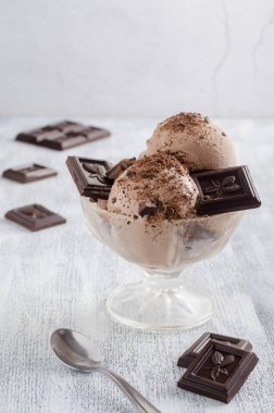 Çikolata parçalı dondurma ve çikolata parçacıkları şeffaf bir kâse içinde beyaz ahşap bir masada, çikolata parçalarının arka planında..