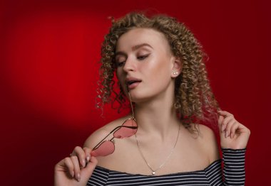 Sarı kıvırcık saçlı, elinde pembe gözlüklü genç bir kız. 