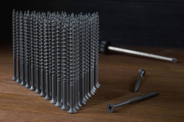 Gri kendi kendine dokunan vidalar ahşap bir masanın üzerinde üçgen şeklindedir, yakınlarda birkaç vida vardır, arka planda bir tornavida vardır..