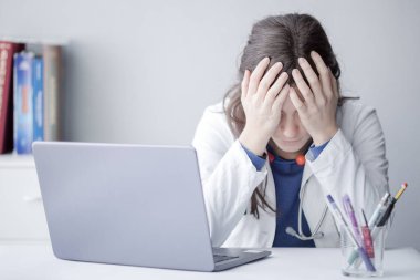 Klinikte dizüstü bilgisayarla çalışan yorgun genç kadın doktor.