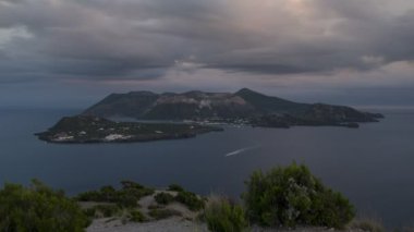 İtalyan adası Lipari 'nin zaman atlaması ön planda ve gün batımında uzakta Vulcano adası, denizde hareket eden tekneler, gökyüzünde bulutlar Sicilya Liparic Adaları