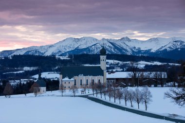 Kış gündoğumunda Wilparting Irschenberg 'deki hac Kilisesi' nde kar, arka planda dağlar, ağaçlı yollar, Bavyera Almanya