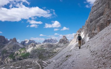 Sırt çantalı bir adam, Avrupa Dolomit Alpleri 'ndeki Three Peaks doğa koruma alanında, dağlık Paternkofel' in kayalık duvarlarının altında yürüyüş yapıyor. Bulutlu mavi gökyüzü, Güney Tyrol İtalya.