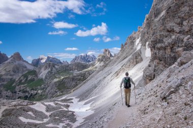 Sırt çantalı bir adam, Avrupa Dolomit Alpleri 'ndeki Three Peaks doğa koruma alanında, dağlık Paternkofel' in kayalık duvarlarının altında yürüyüş yapıyor. Bulutlu mavi gökyüzü, Güney Tyrol İtalya.
