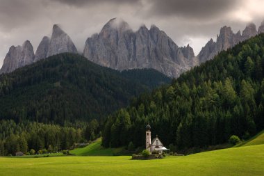 St. Johann kilisesi St. Magdalena 'da dağ manzarası, yeşil çayır üzerinde güneş, arka planda orman ve dağlar, gökyüzünde ağır bulutlar, Val Di Funes, Dolomite Alpleri, Güney Tyrol İtalya