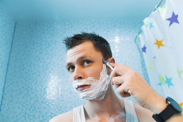 A man shaves his face in the foam with a razor. Guy shaves in front of a mirror