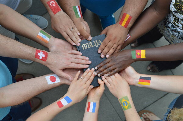 International brothers and sisters in Christ with different flags painted on their arms holding a bible together