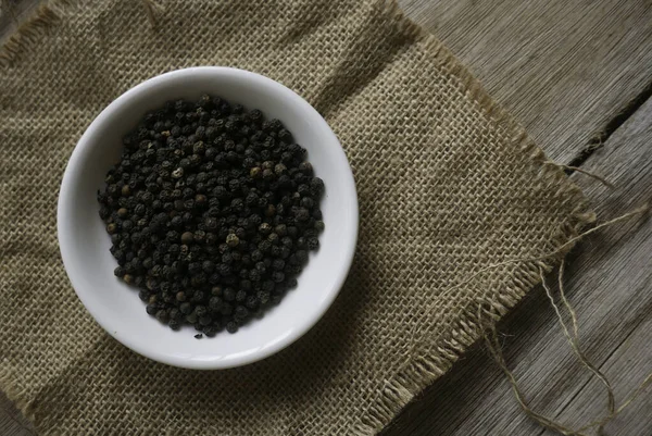 Pimienta negra en un plato blanco en un saco de alfombra sobre fondo de madera . — Foto de Stock