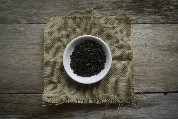 Pimienta negra en un plato blanco en un saco de alfombra sobre fondo de madera . — Foto de Stock