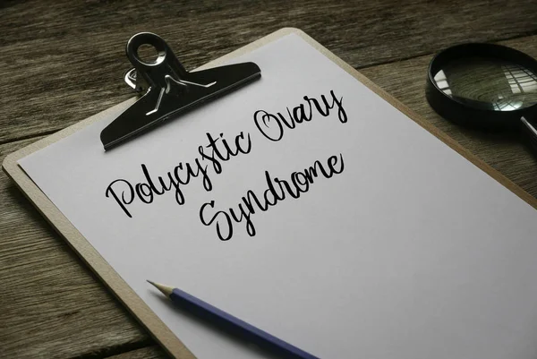 Kaca pembesar, pensil dan papan klip dengan kertas putih yang ditulis dengan Polycystic Ovary Syndrome di latar belakang kayu . Stok Gambar Bebas Royalti