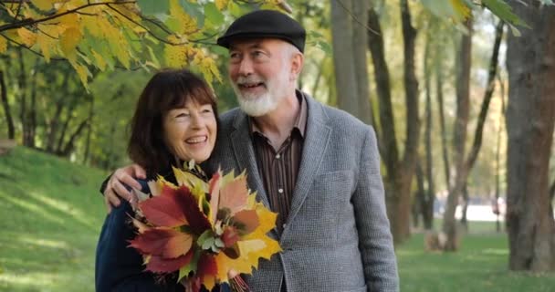 高齢者のカップルは、木々の間の居心地の良い公園で幸せを笑い、抱きしめ、晴れた秋の日を楽しむ-スローモーション。引退後の現役現代生活。幸せと笑顔. — ストック動画