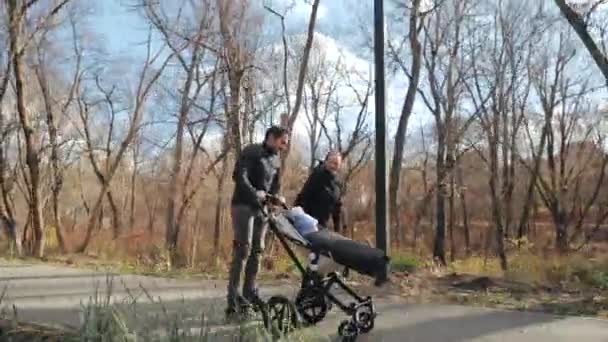 Сімейні виступи в осінньому парку, роликових лопатях і колясці. Тато наздоганяє маму і дає їй коляску. Батьки на валиках з дитиною в повільному русі . — стокове відео