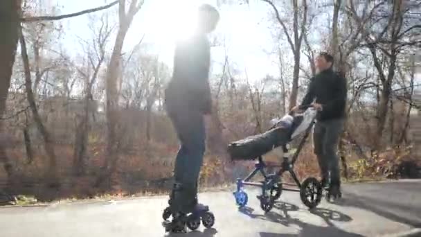 Patines deportivos de la familia joven con un bebé en un cochecito en un cálido parque de otoño. Mamá cabalga hacia atrás y sonríe. En cámara lenta. Educación deportiva y pasión saludable . — Vídeo de stock