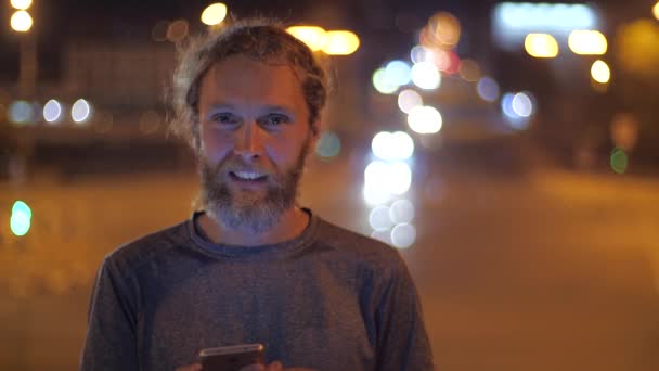 Закріпіть Портрет вродливого кавказького бородатого молодого чоловіка з довгим волоссям і смартфоном, який запрошує когось жестами і мімімікою. Переміщення розжарених вечірніх вогнів міста на задньому плані — стокове відео