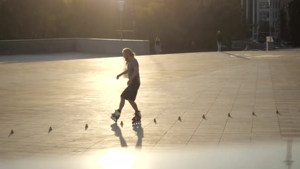 Молодий довгошерстий чоловік-ролик танцює між конусами ввечері на міській площі на заході сонця. Фрістайл слалом катання на ковзанах між конусами в повільному русі . — стокове відео
