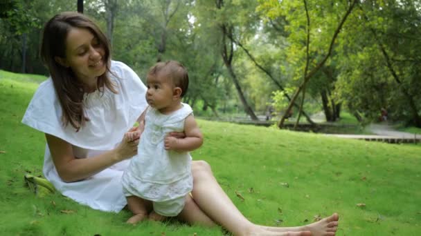 Giovane bella madre in un abito bianco insegna alla figlia appena nata a camminare su un'erba verde fresca in un parco. Felice giovane madre e bambino imparare a camminare all'aperto e godersi la natura . — Video Stock