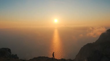 Silhouette of a Young Woman walking along a cliff edge observing a beautiful dramatic sunset above a sea from a high mountain in Crimea. Lady hiker with arms outstretched against beautiful sunset