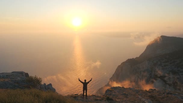 Silueta de una mujer joven de pie en un borde del acantilado observando una hermosa puesta de sol dramática sobre un mar desde una alta montaña en Crimea. Senderista con los brazos extendidos contra la hermosa luz del atardecer . — Vídeo de stock