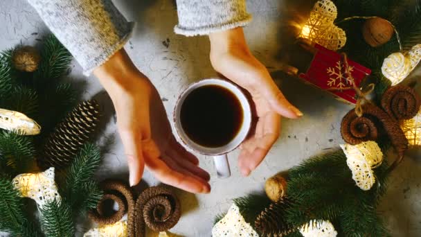 Female hands slowly pick up a cup of tea or coffe from the table. Girl warms her hands over a hot cup. Nearby are Christmas decorations, spruce branches, decorative sledges, garlands in form of hearts — ストック動画