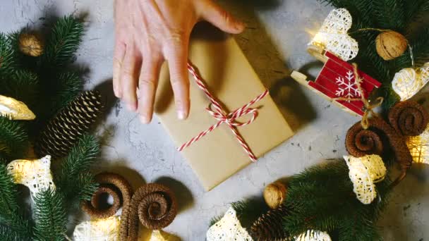 Πρωτοχρονιάτικο δώρο ανάμεσα σε έλατο και πεύκα, κώνους από μίνι έλκηθρα, ξηρούς καρπούς και γιρλάντα. Το αρσενικό χέρι σηκώνεται αργά από το τραπέζι. Χειμερινές διακοπές πλησιάζουν, Πρωτοχρονιά και Χριστούγεννα — Αρχείο Βίντεο