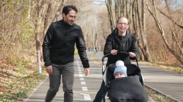 Jonge gelukkige en atletische familie glimlacht en geniet van rolschaatsen met hun kind in een wandelwagen langs de zonnige steeg van het herfstpark. Papa knipoogt naar het kind. Langzame beweging. — Stockvideo
