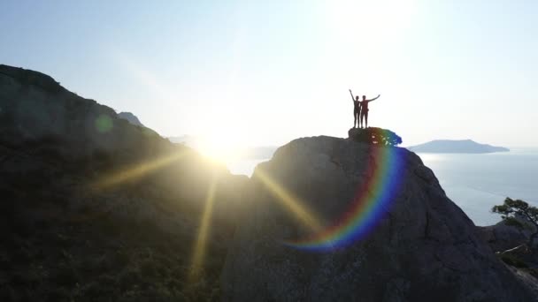 Lotnicza sylwetka kochającej się pary stojącej na szczycie góry nad morzem o wschodzie słońca i machającej rękami. Lot nad młodymi aktywnymi alpinistami mężczyzna i kobieta na szczycie na Krymie — Wideo stockowe