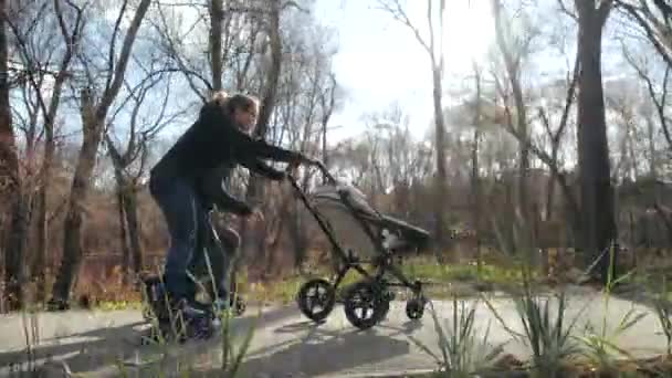 Szczęśliwa młoda rodzina jeżdżąca na wrotkach ze swoim dzieckiem w wózku w parku miejskim. Tata robi niezwykłe akrobacje na wrotkach i upadkach. Jasna, słoneczna jesień.. Zwolniony ruch. — Wideo stockowe
