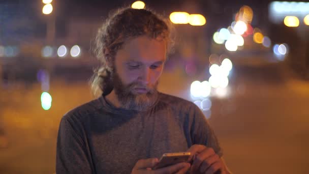 Close-up, portret van een knappe blanke bebaarde langharige jonge man die smartphone gebruikt op de achtergrond van het verplaatsen van Avondverlichting van een stad. — Stockvideo