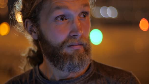 Retrato de cerca de un guapo caucásico barbudo joven de pelo largo mirando con interés y asombro el fondo de las luces de la noche en movimiento de una ciudad . — Vídeos de Stock