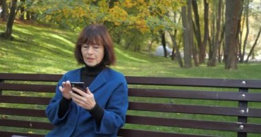 Sonbahar parkında otururken telefon mesajı yazan yaşlı bir kadın. Akıllı telefondan ekrana bakan mutlu yaşlı kadın. Ağır çekimde akıllı telefonda kablosuz internet bağlantısı kullanan yaşlı bir kadın..