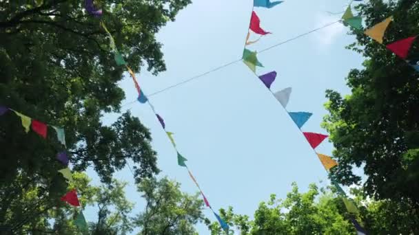 Ünnepi koszorú többszínű papír zászlók az ünnep. Tájkép egy gyerekzsúrra. Természeti dekoráció a fák hátterében. Bunting banner - színes zászlók A lapok. — Stock videók